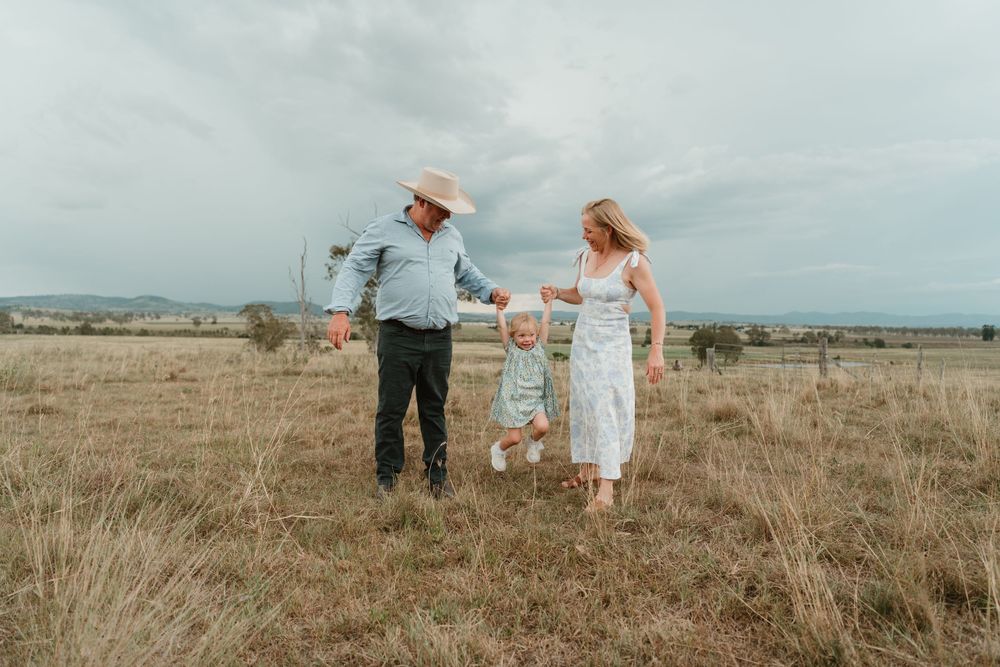 Family of three swinging toddler in grassy field, parents holding hands with child under cloudy rural sky.