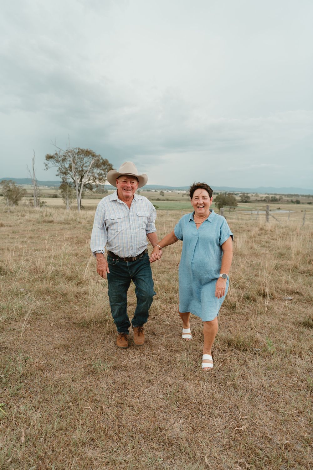 Elderly couple holding hands and smiling while walking through a dry rural field under a cloudy sky.