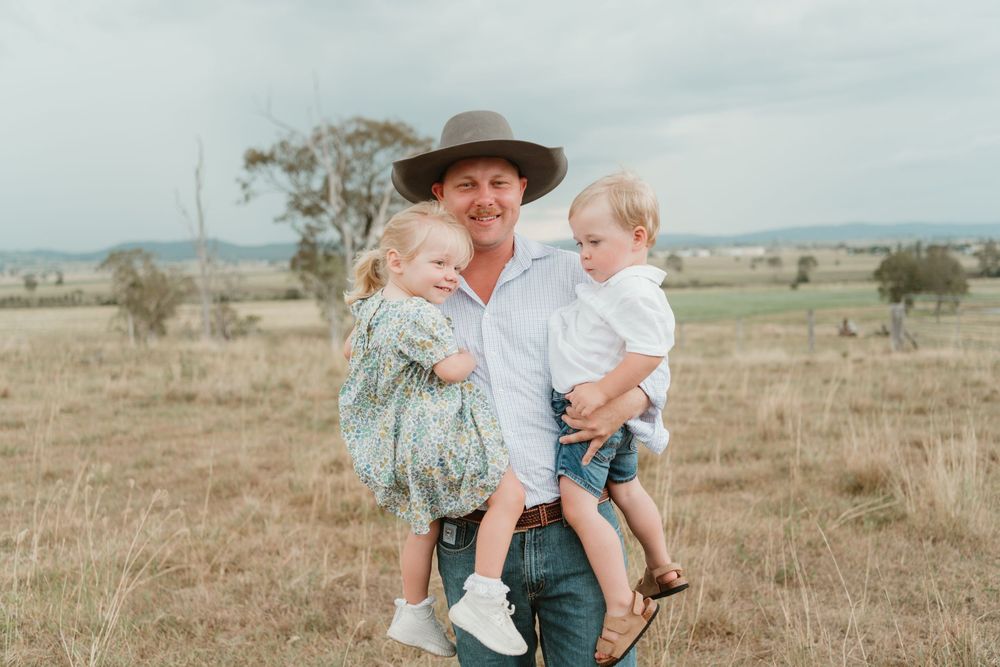 A man in a wide-brim hat holds two young children in a rural field with dry grass and hills in the background.