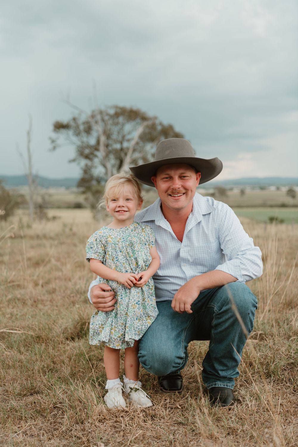 A smiling farmer in a wide-brim hat crouches beside a young blonde girl in a floral dress in a rural field.