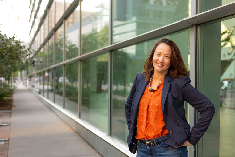 Professional wearing navy blazer and orange blouse poses confidently against modern glass building exterior.
