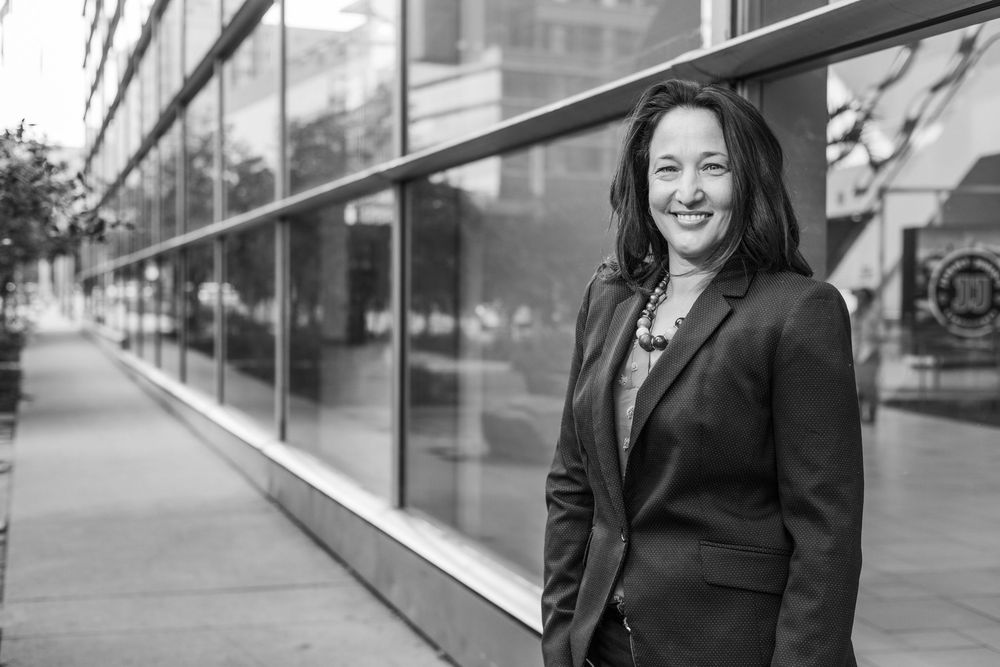 Professional business figures walk alongside modern glass office building in urban setting while smiling.