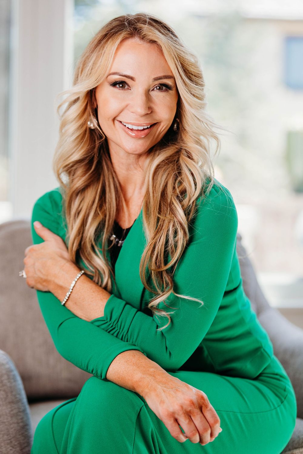 Professional portrait of someone in a vibrant emerald green outfit sitting on a gray couch with a bright smile.