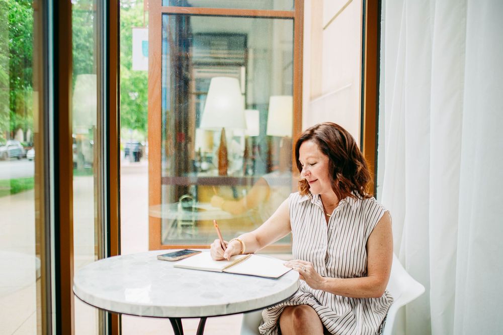 Interior view of a casual coworking space with bright sunlit windows and a person working at a round marble table.