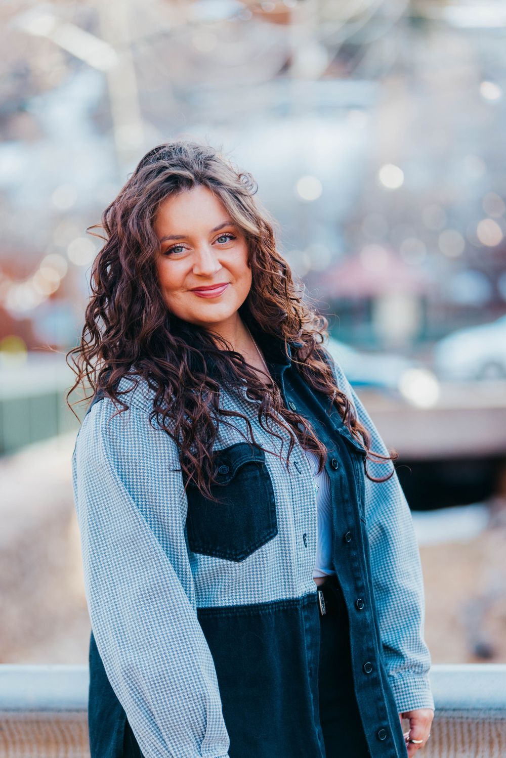 Woman in denim jacket with curly dark hair poses against blurred background.