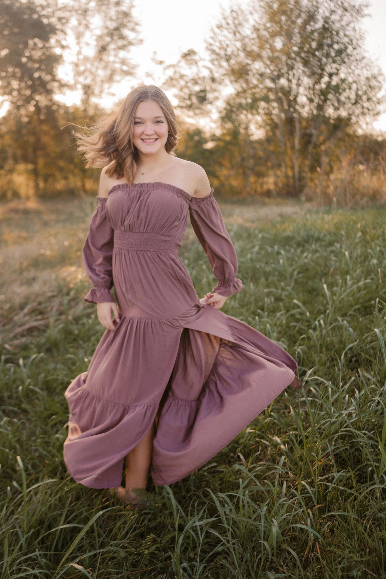 A flowing mauve off-shoulder dress twirls gracefully in a golden sunlit field of tall grass at sunset.