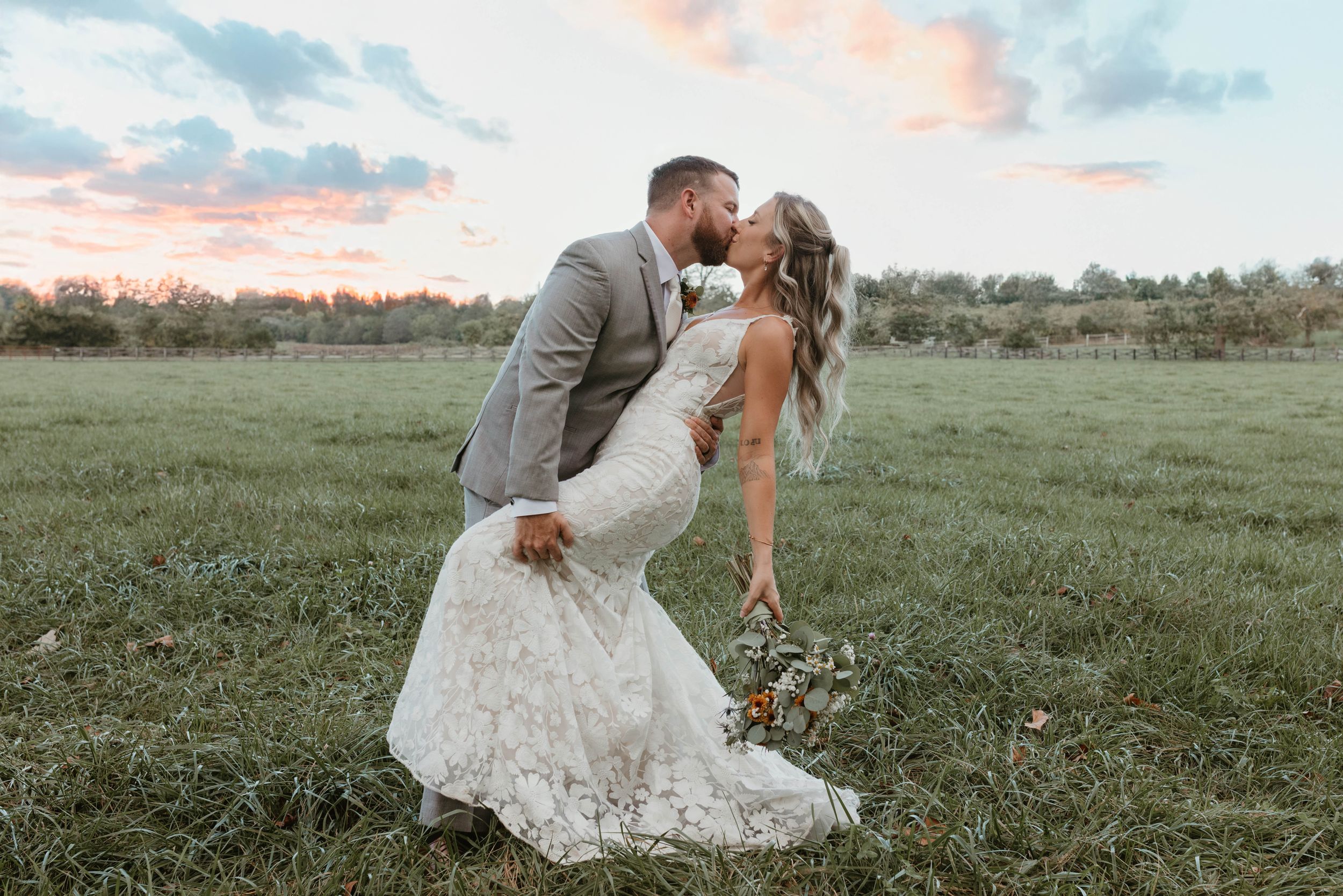 A couple in wedding attire poses in a meadow during golden hour.