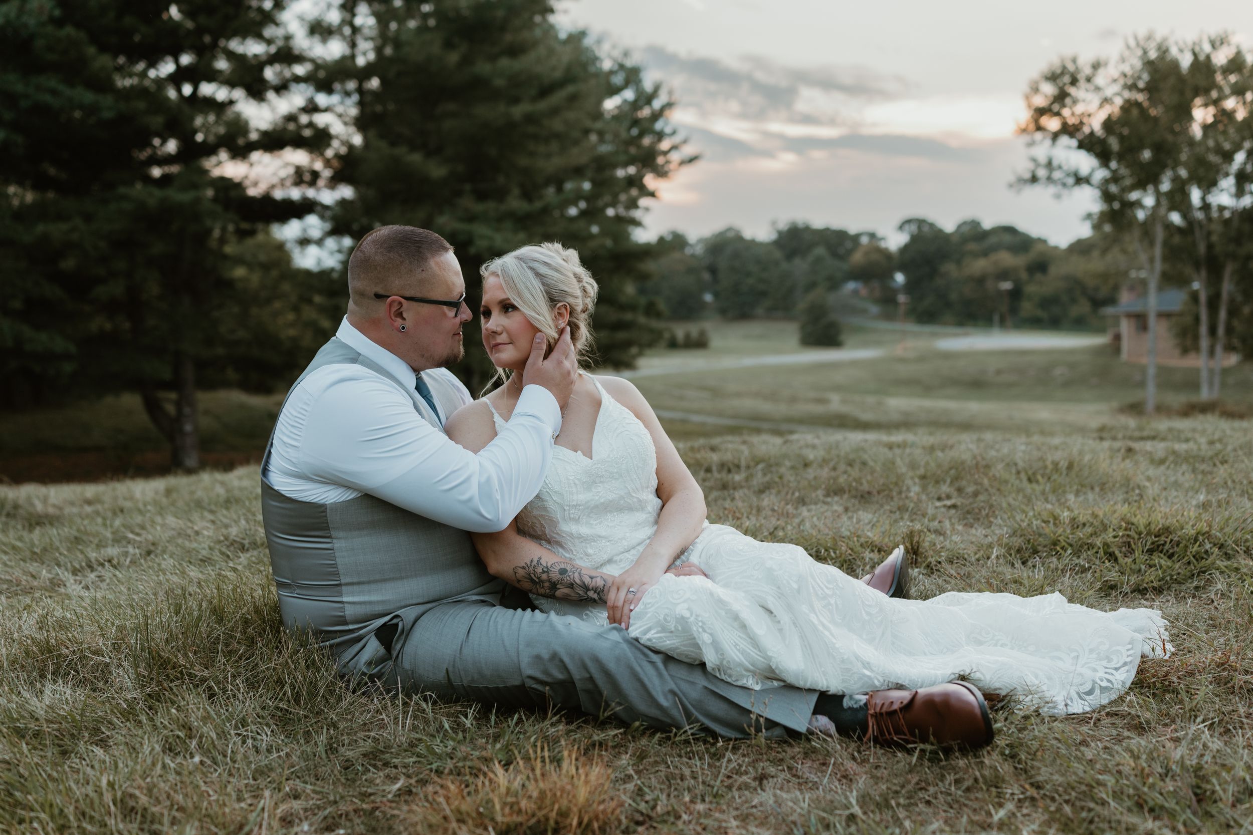 Wedding couple shares intimate moment while sitting on grassy hill at dusk.