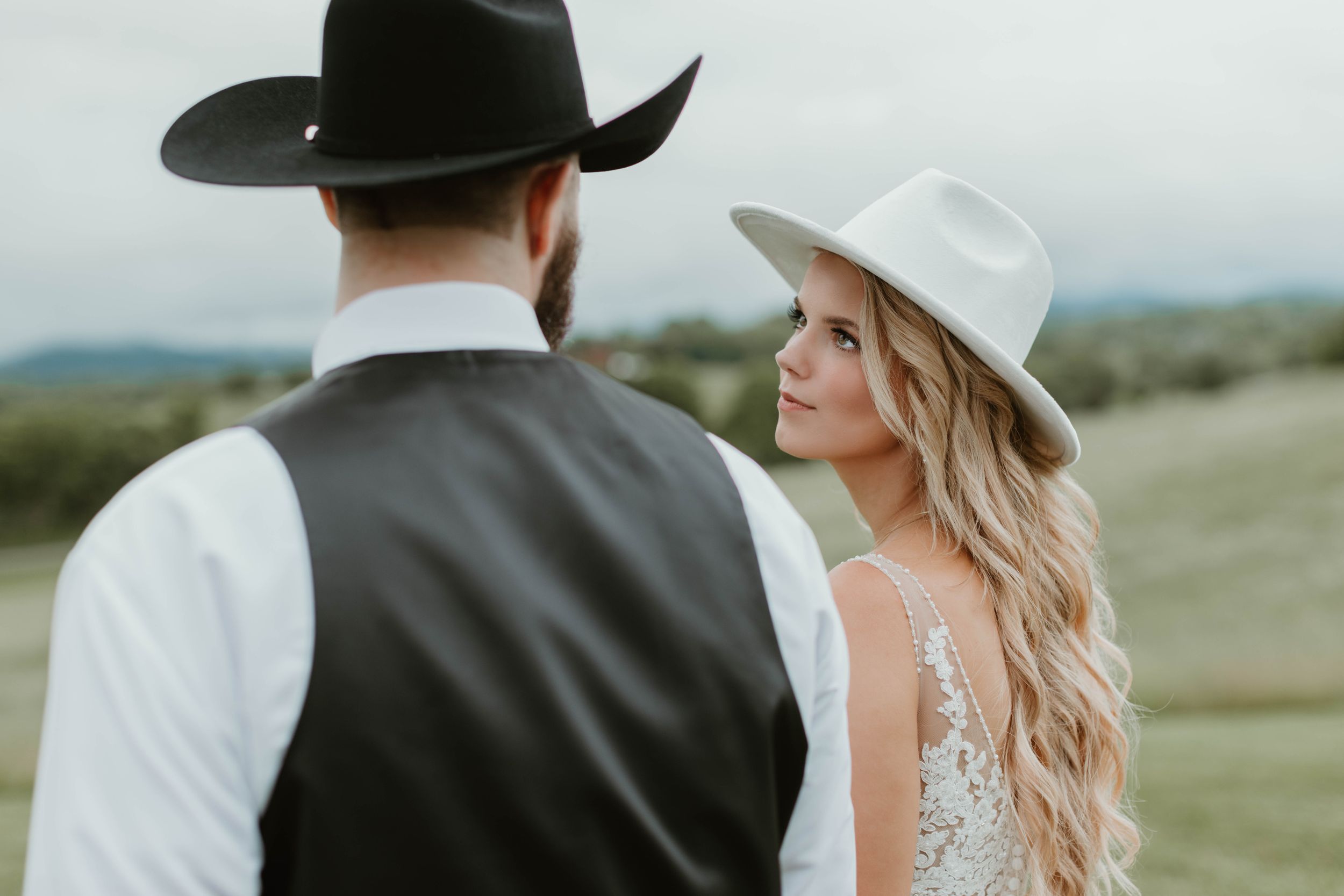 Romantic outdoor wedding portrait with cowboy hats against a scenic landscape.