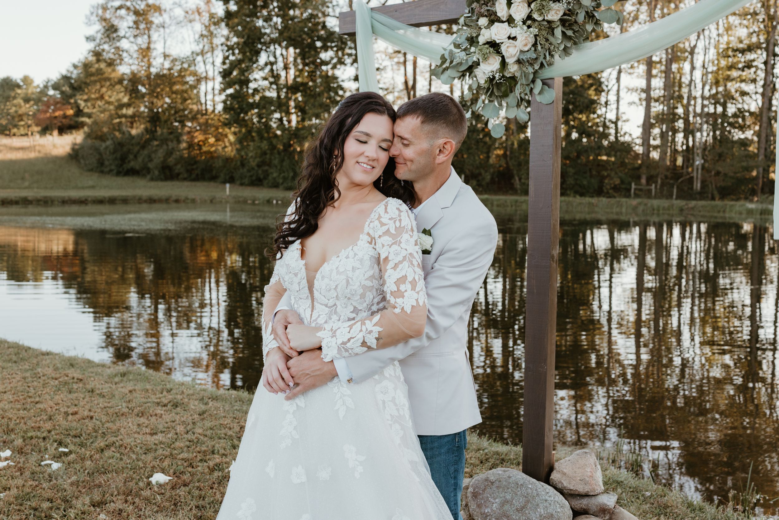 Romantic wedding couple embraces by lakeside under decorated arch at sunset.