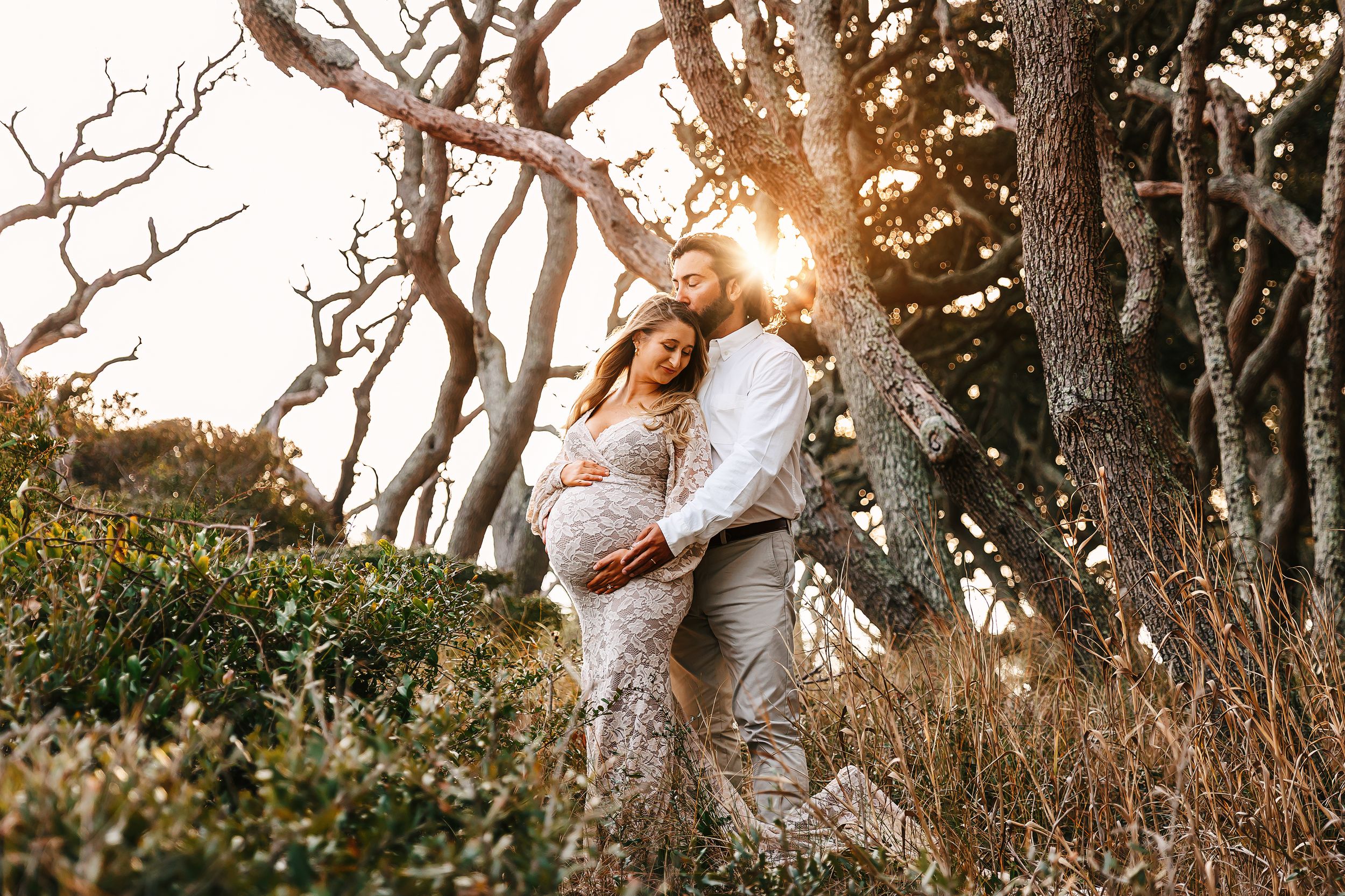 Couple embraces in a romantic sunset moment among bare winter trees and tall grass at golden hour.