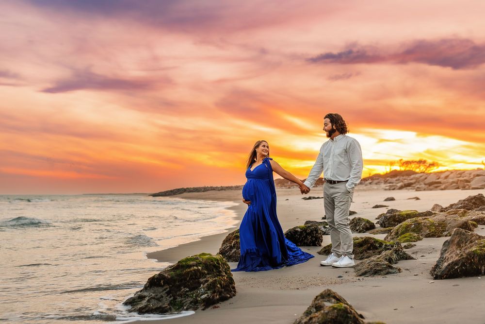 A couple in formal attire stands on rocky coastal cliffs during a dramatic orange and purple sunset.