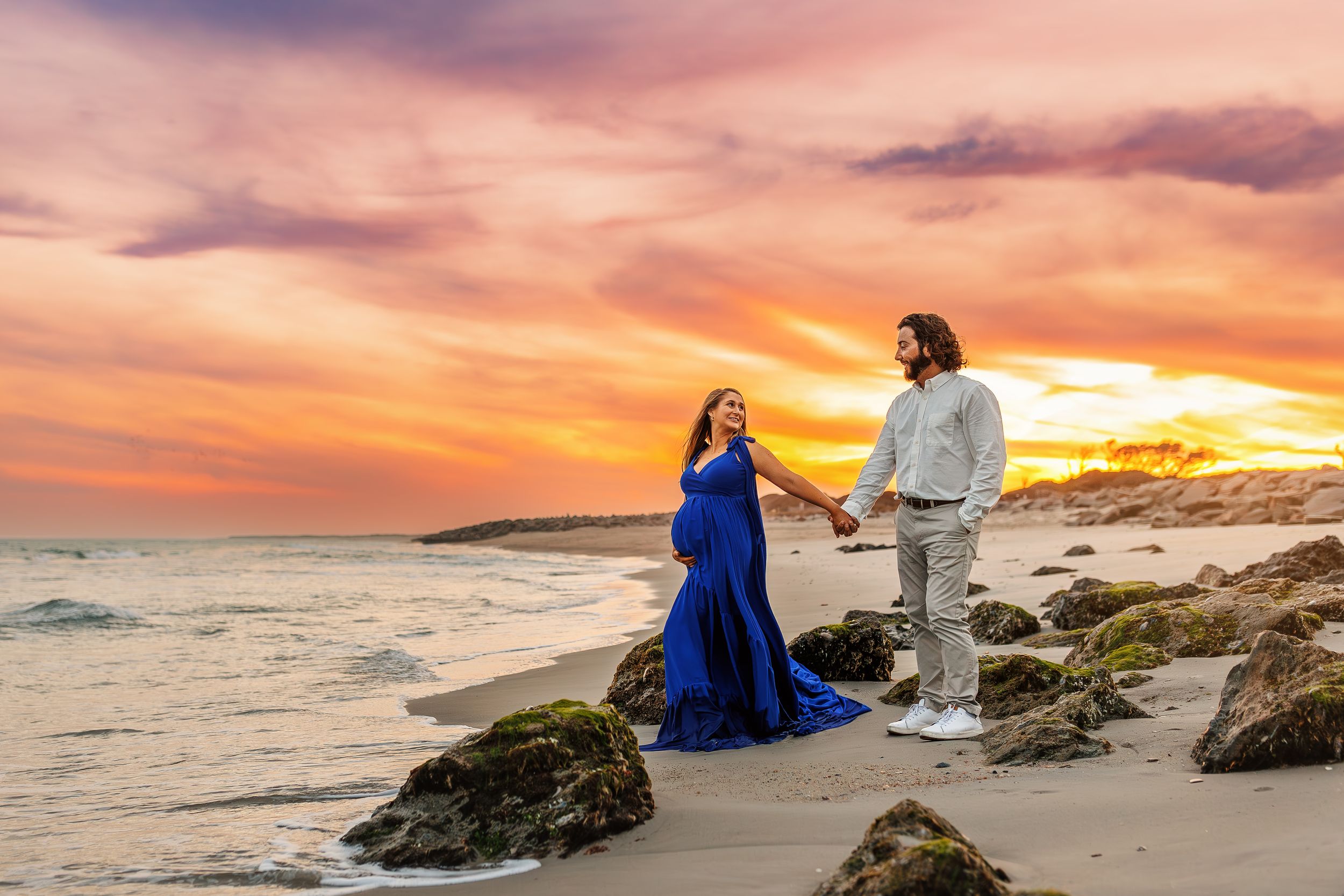 A couple in formal attire stands on rocky coastal cliffs during a dramatic orange and purple sunset.