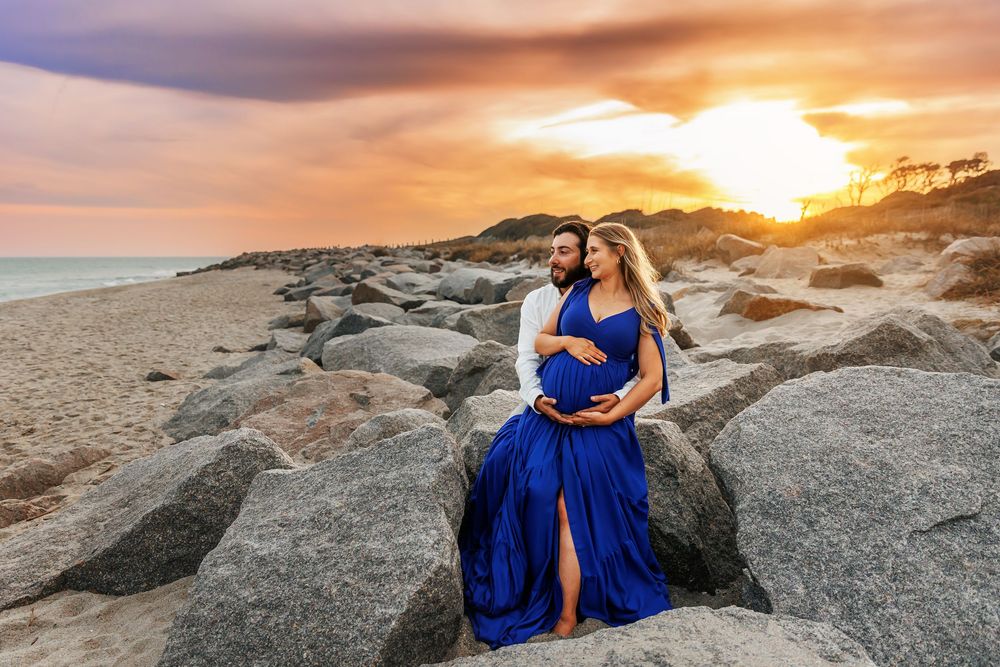 A figure in a flowing royal blue gown poses gracefully on coastal rocks at sunset creating a dramatic silhouette.
