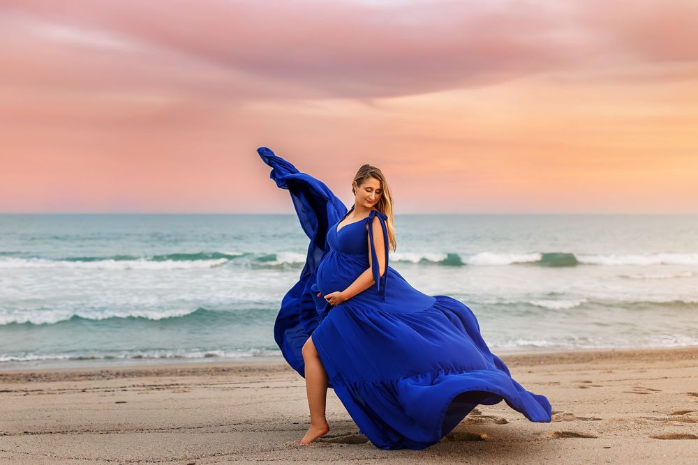 A person in a flowing royal blue dress poses gracefully on a sandy beach at sunset with ocean waves in the background.
