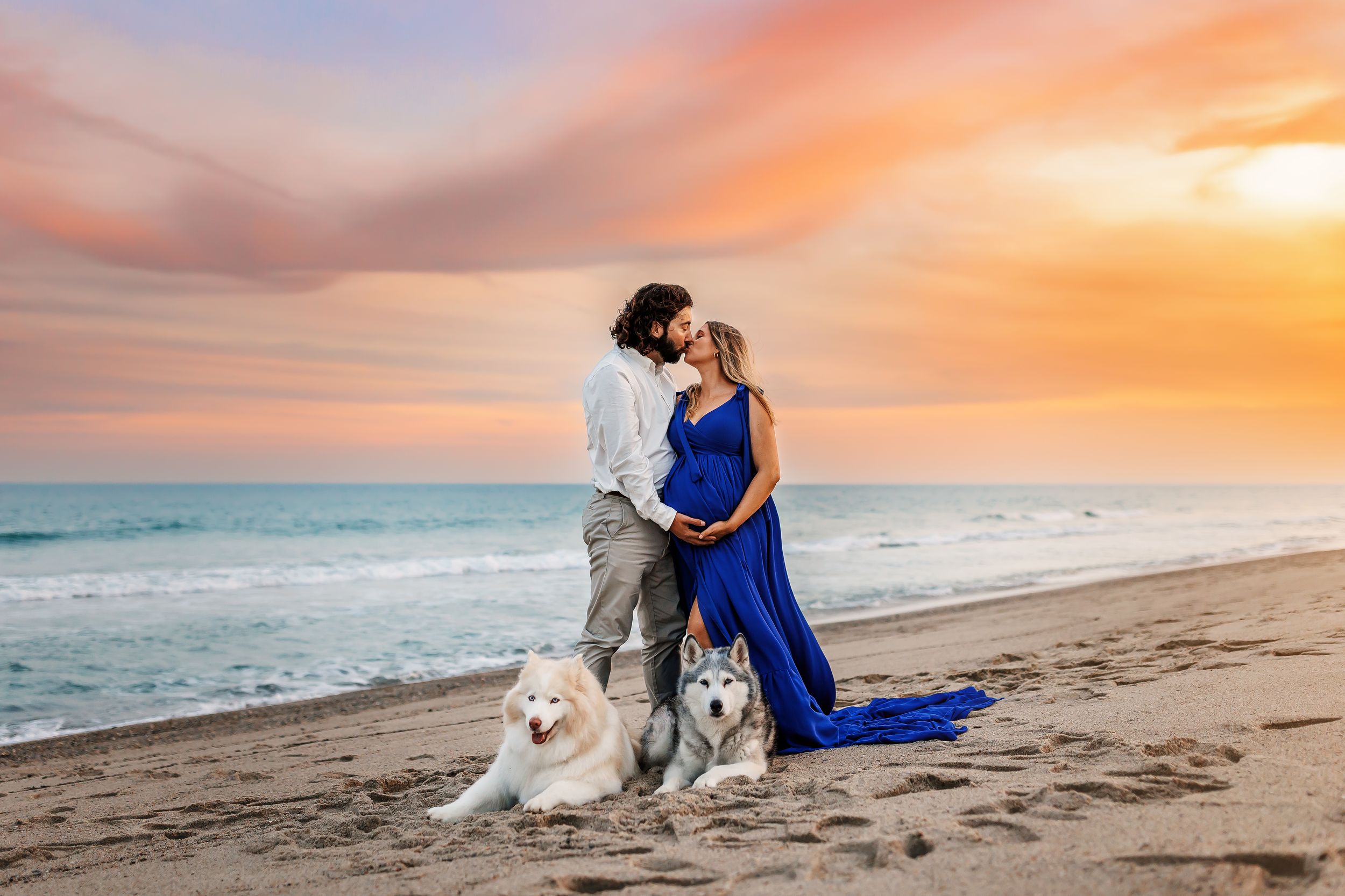 A romantic beach photoshoot at sunset with a white dog sitting in the sand beside a couple in formal attire.