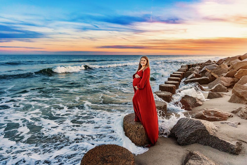 Woman in red dress poses dramatically on rocky shoreline during golden hour sunset.