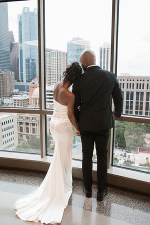 A couple in wedding attire gazes out floor-to-ceiling windows at a cityscape view of tall buildings and skyscrapers. Houston wedding