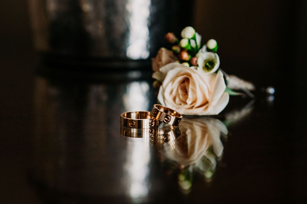Wedding rings and a light pink rose with greenery sit on reflective dark surface with bokeh effect in background.