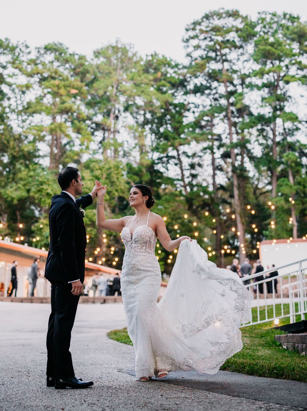 A couple dancing on a patio surrounded by string lights and tall trees at dusk during their wedding celebration.
