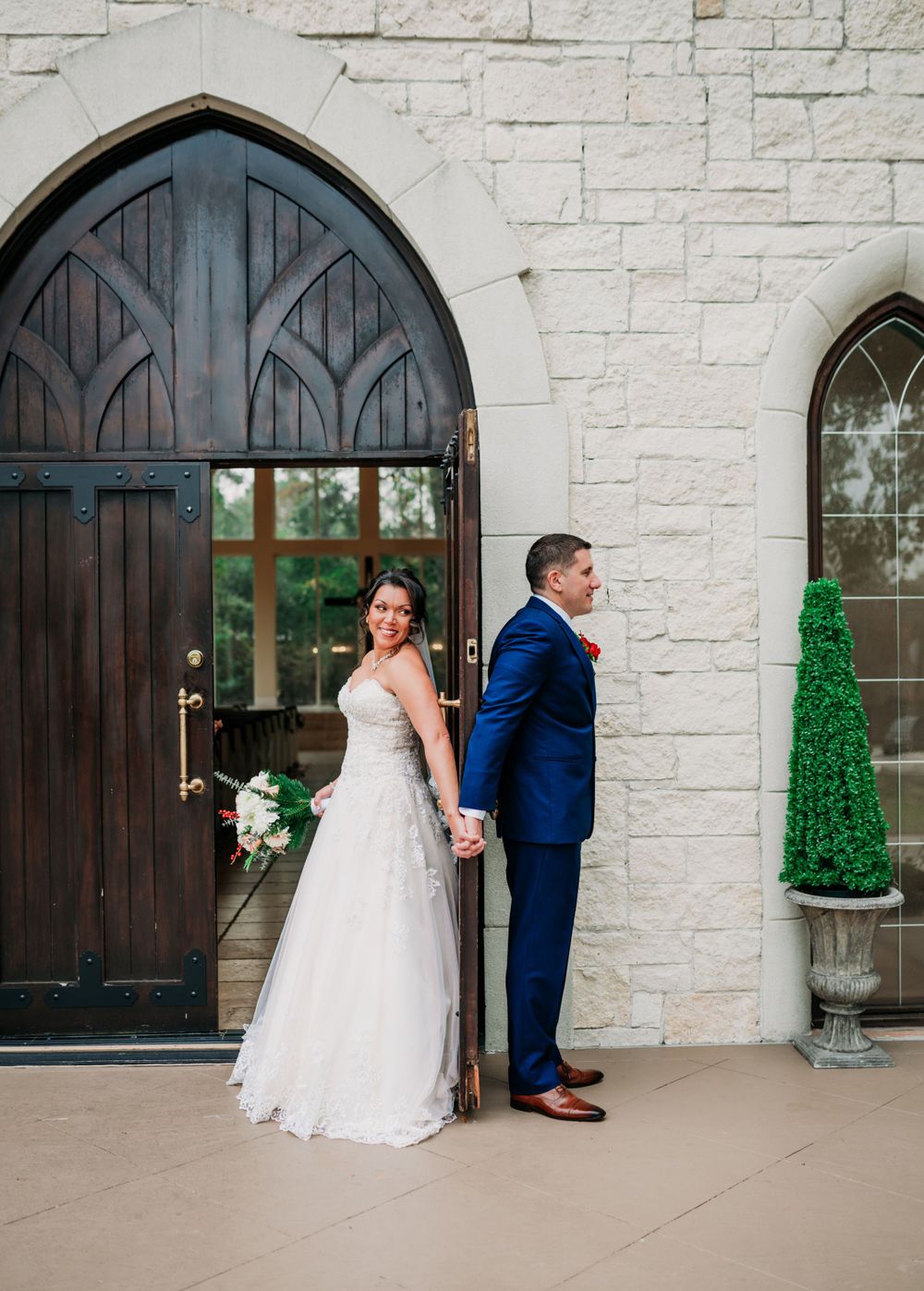 A couple poses back-to-back for a wedding photo in front of a grand wooden arched door and white stone church facade.