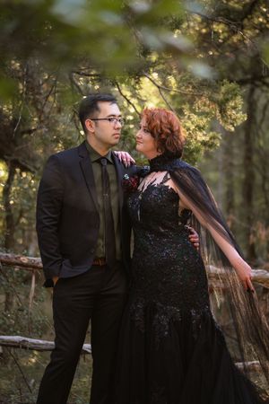 A couple in formal attire share an intimate moment together in a sunlit wooded setting during a romantic photoshoot. Halloween Wedding