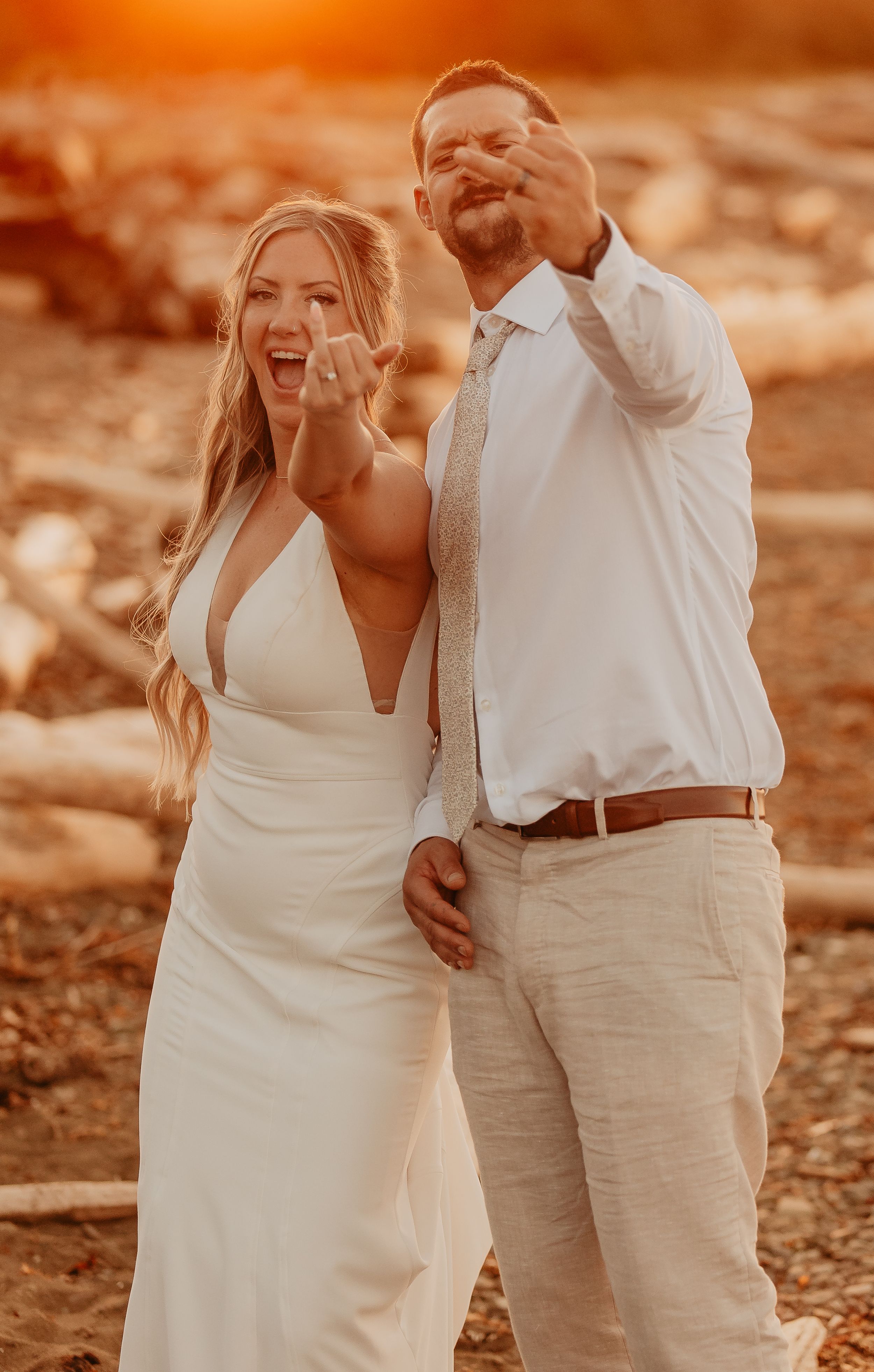 A couple in white attire shares candid laughter during a sunset photoshoot with warm orange tones.