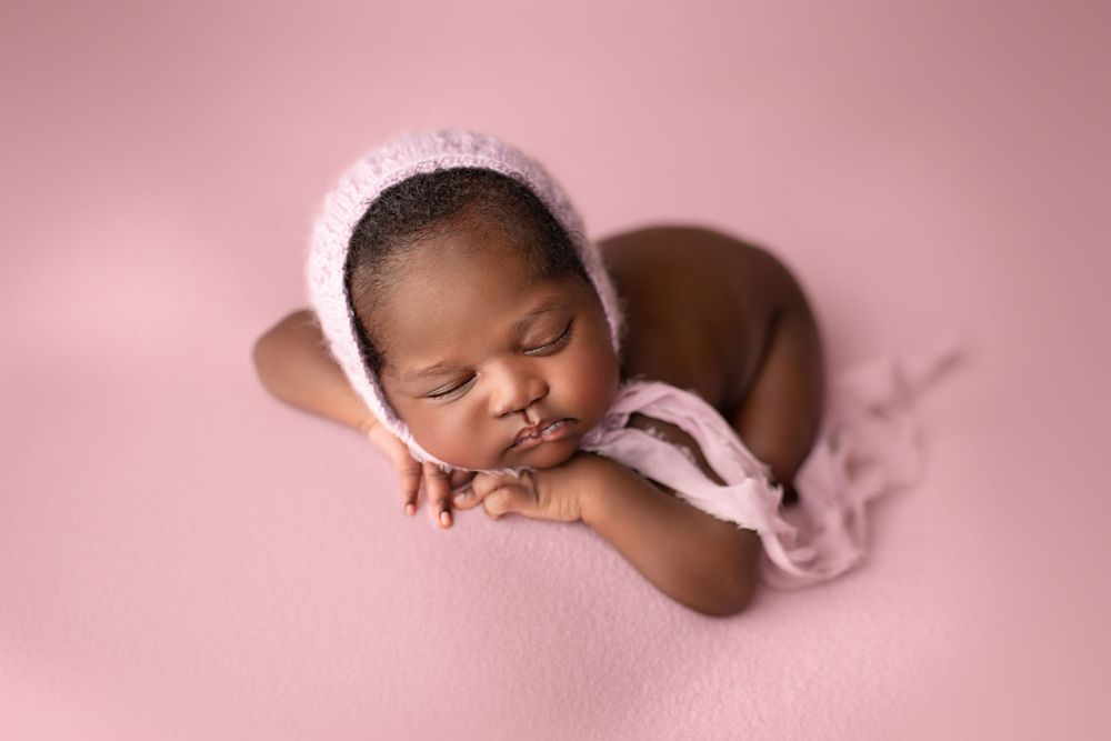 Affordable newborn photographer Springfield Lakes – African baby girl in white bonnet sleeping curled on pink backdrop, professional baby po