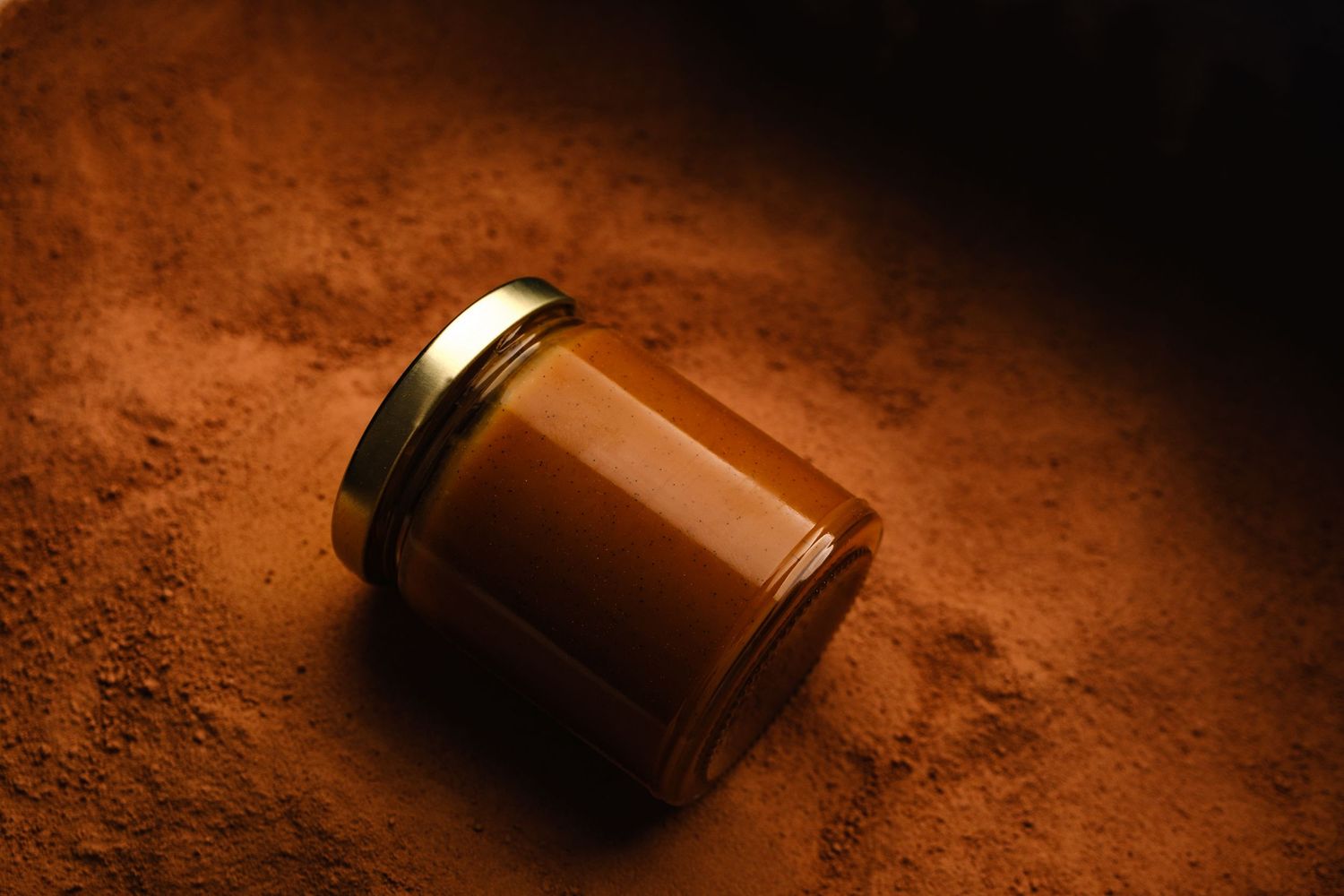 Glass jar of caramel spread lying on its side on a bed of brown powder in warm light.