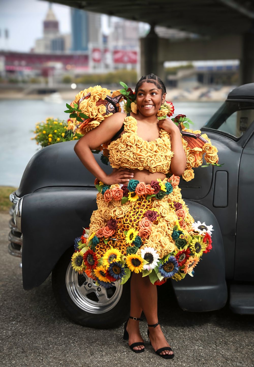 A creative floral dress made entirely of sunflowers and yellow flowers is worn while posing next to a vintage black car.