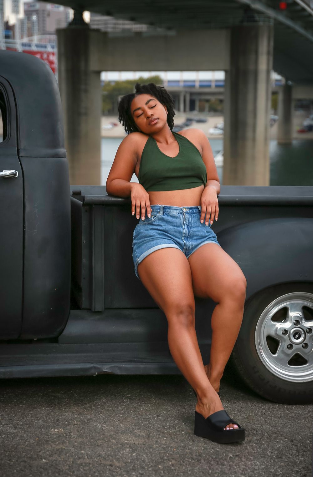 Woman in green crop top and denim shorts poses casually against a black vintage pickup truck in an urban setting.