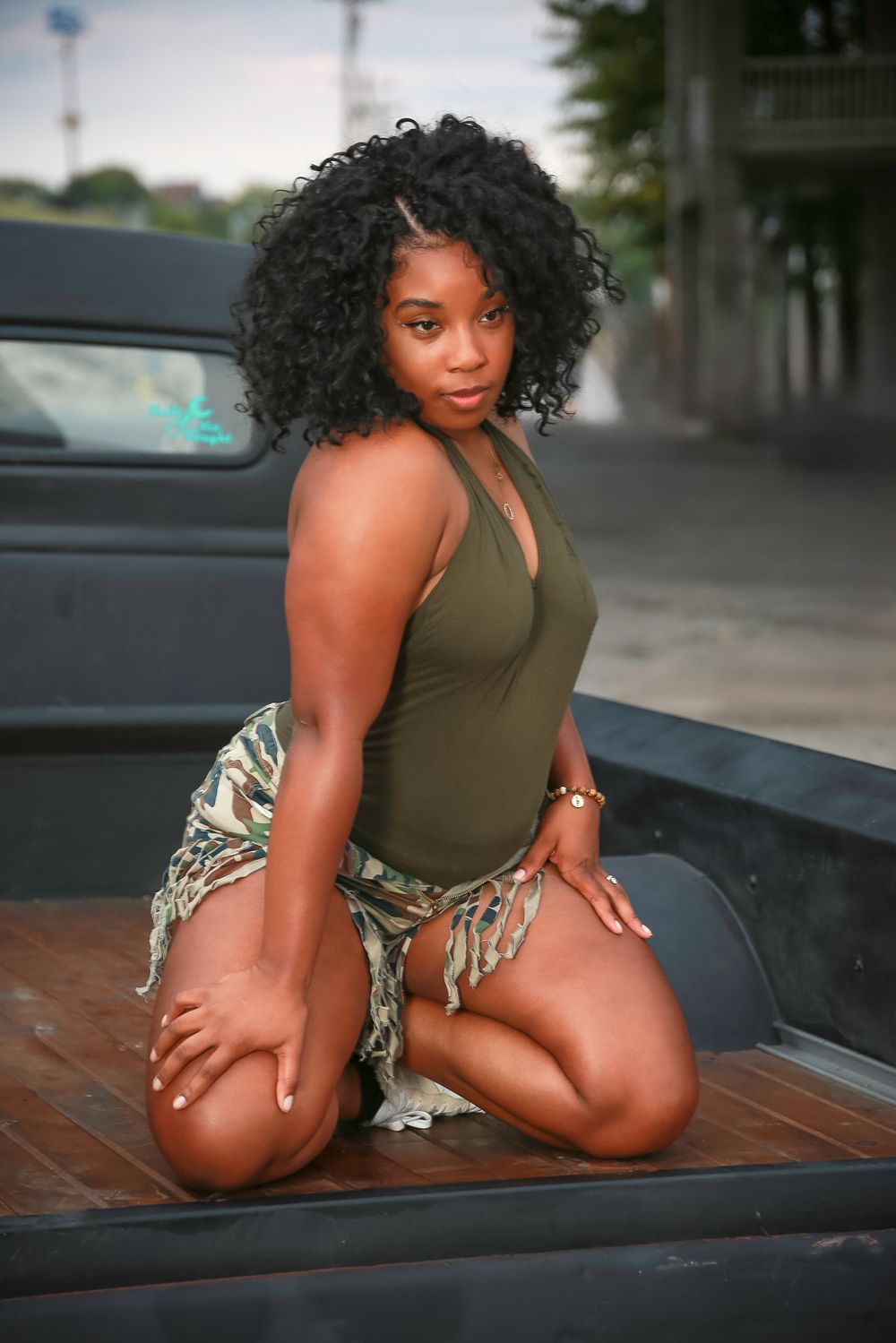 Stylish woman in olive tank top and patterned skirt poses on truck bed during urban photoshoot.