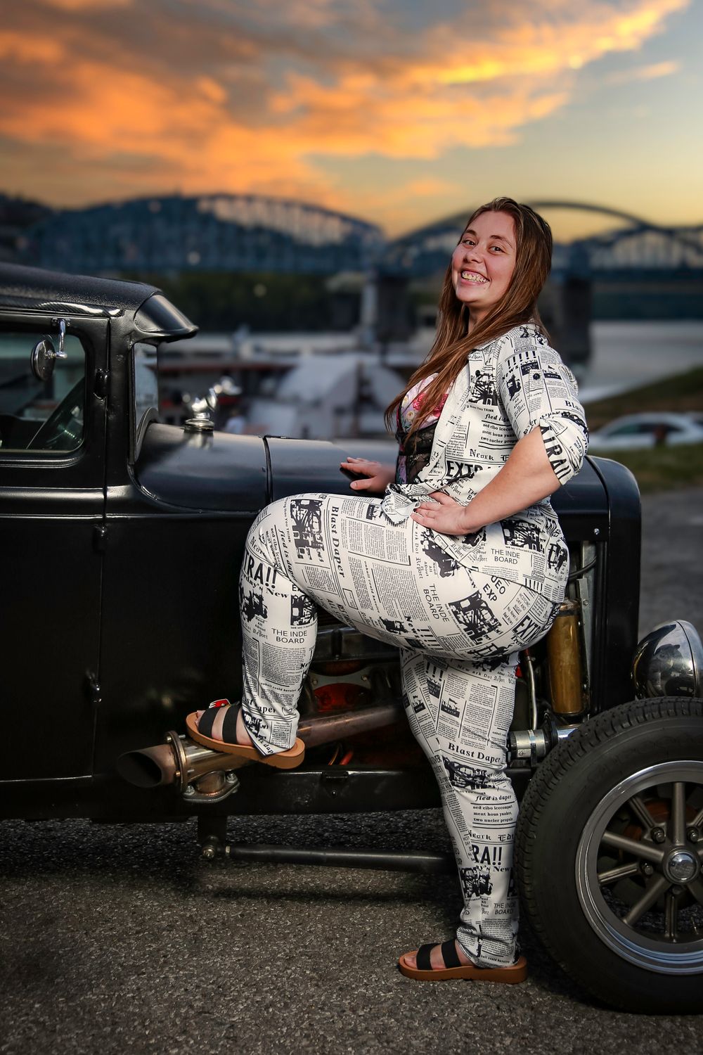 Stylish figure in newspaper print jumpsuit poses with vintage hot rod against dramatic sunset bridge backdrop.