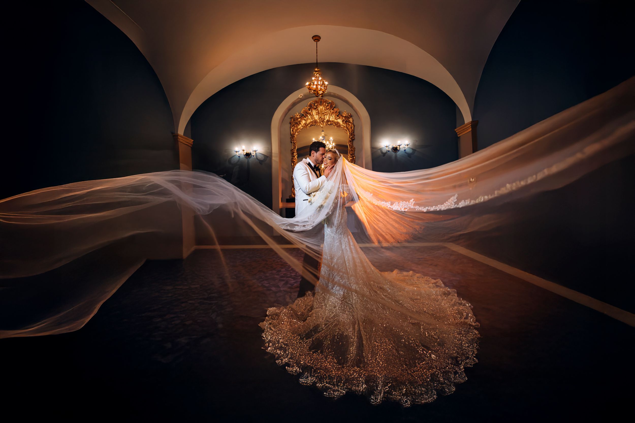Dramatic bridal portrait in arched hallway with flowing veil creating ethereal movement against ornate lace wedding dress.