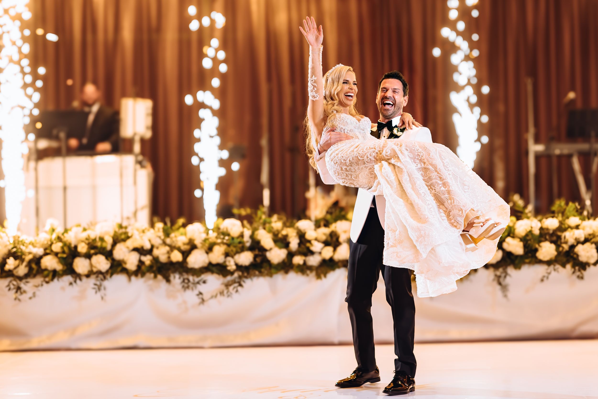 A joyful wedding couple shares a romantic moment on the dance floor with twinkling lights and floral decorations behind them.