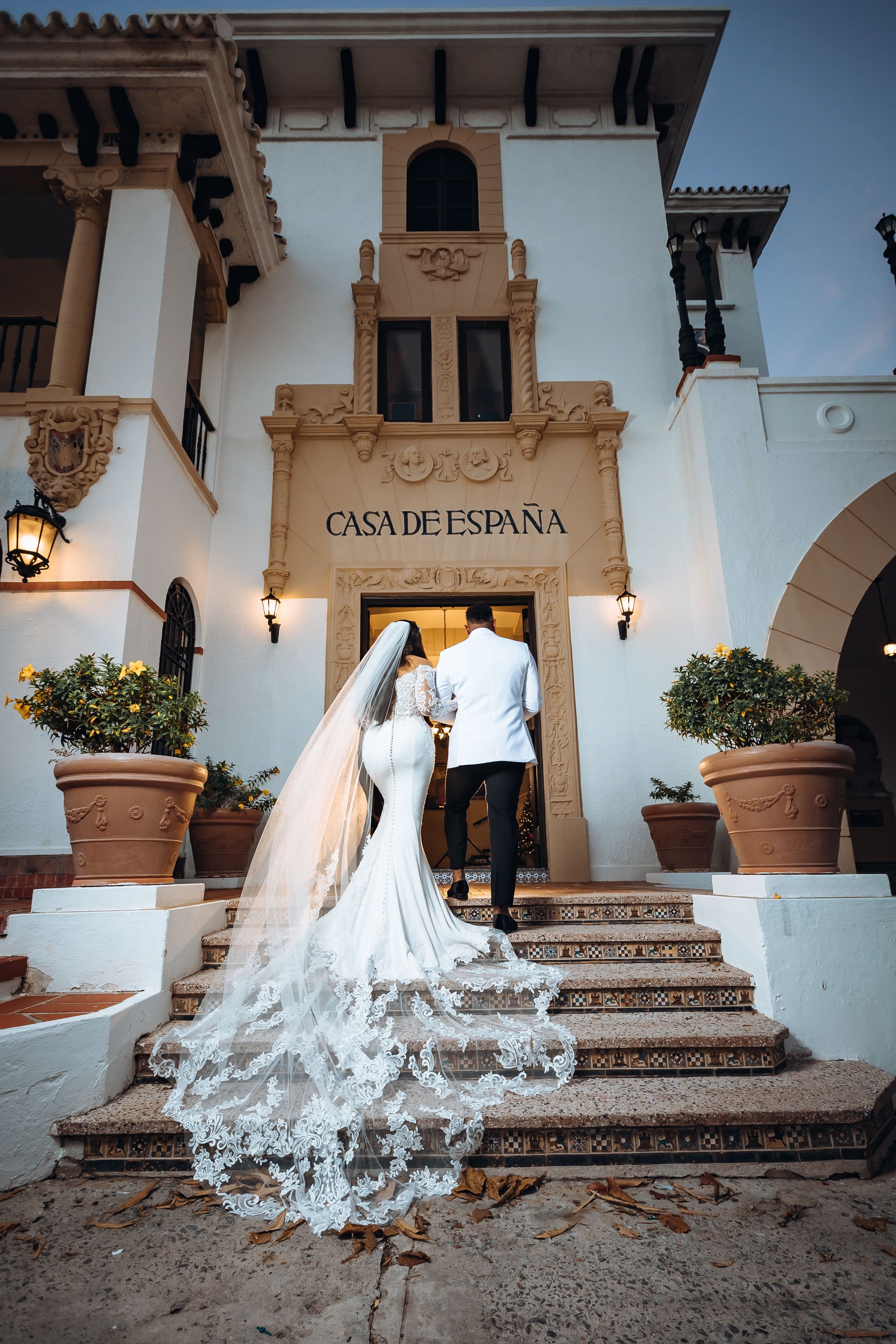 Wedding couple ascending steps of Casa de España with bride's lace train trailing behind.