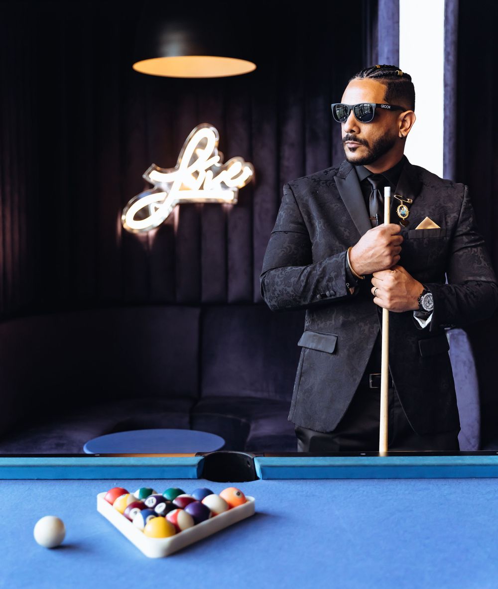 Stylish person in suit and sunglasses poses with pool cue against neon-lit bar backdrop.