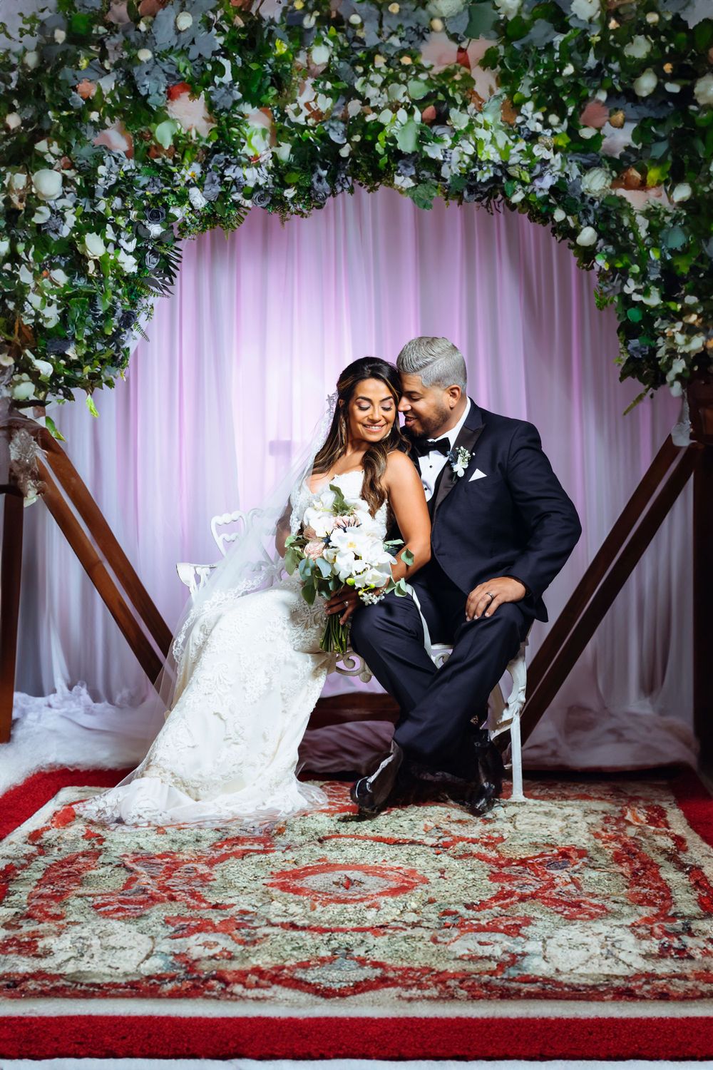 Wedding couple posing under a floral arch with white flowers and pink draping.