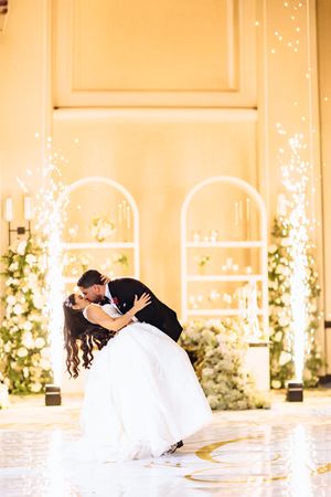 A romantic wedding moment with a dramatic dip kiss in front of illuminated archways and twinkling lights.