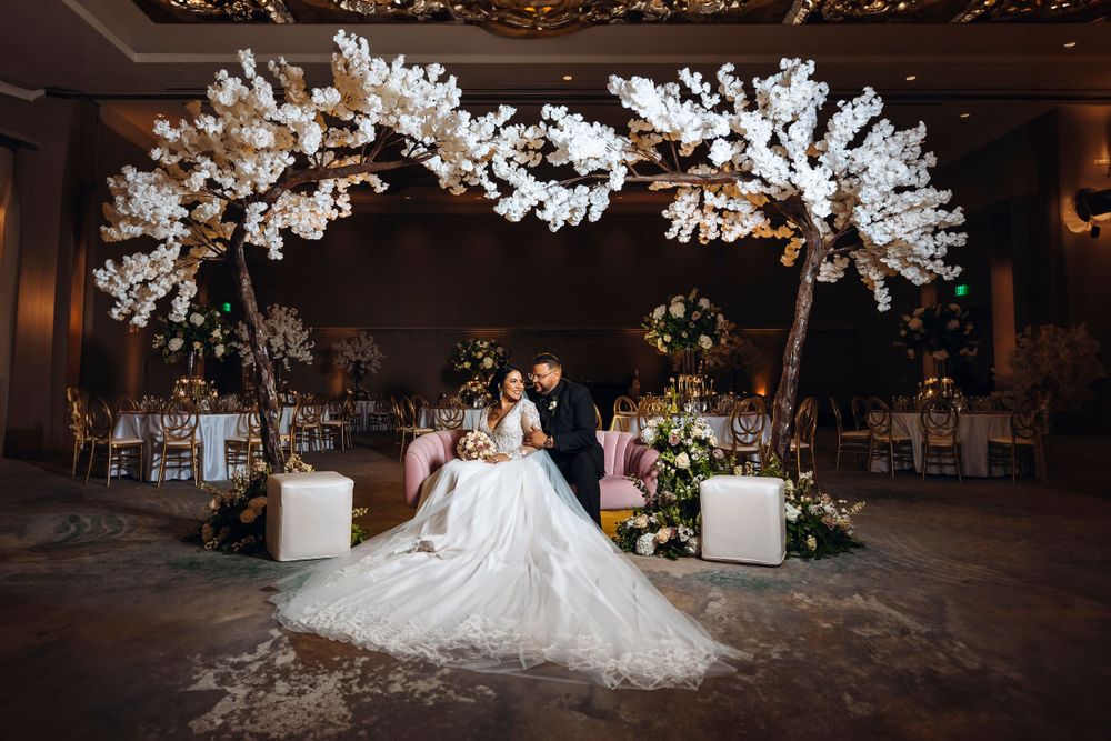 Romantic wedding portrait under illuminated white floral arch in elegant ballroom setting.
