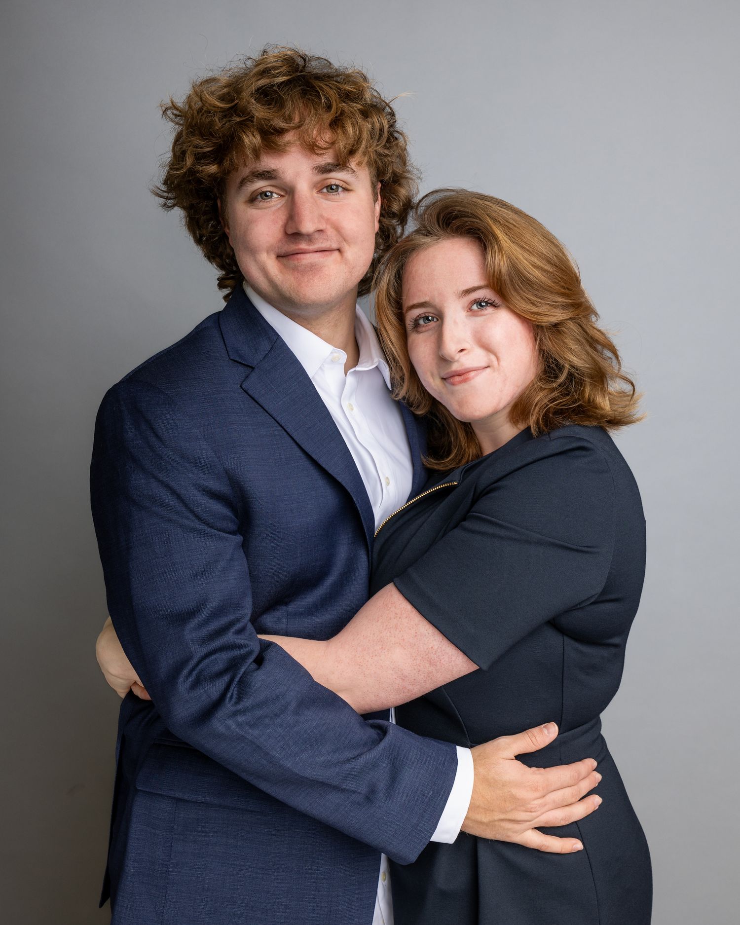 Studio portrait of couple in formal attire embracing against grey background.