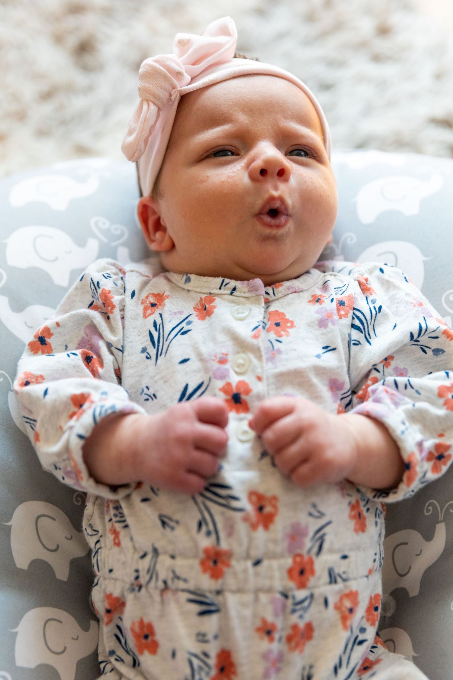 Baby wearing floral outfit and pink bow headband lying on elephant print blanket.