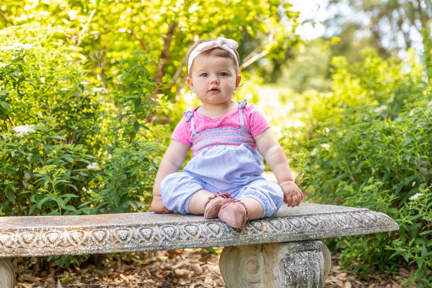 Baby in pink overalls sitting on stone bench surrounded by lush garden foliage.