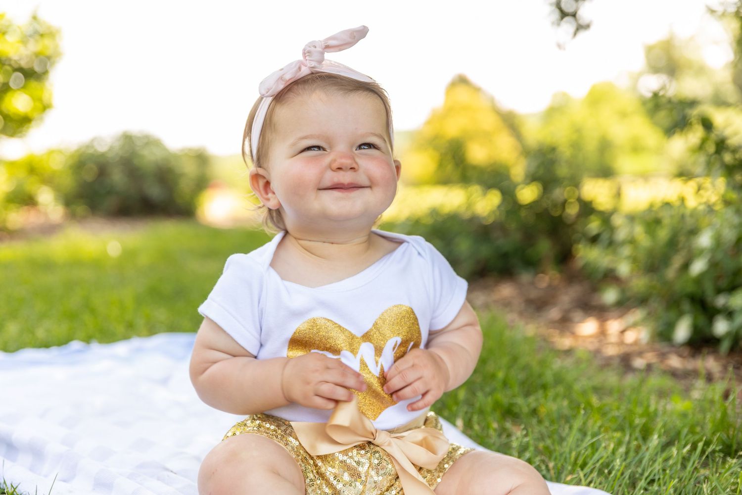 Smiling baby in white top and gold shorts sitting on blanket in sunny garden.