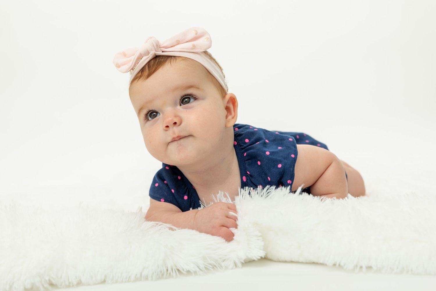 Baby in white headband lying on soft white blanket during studio session.