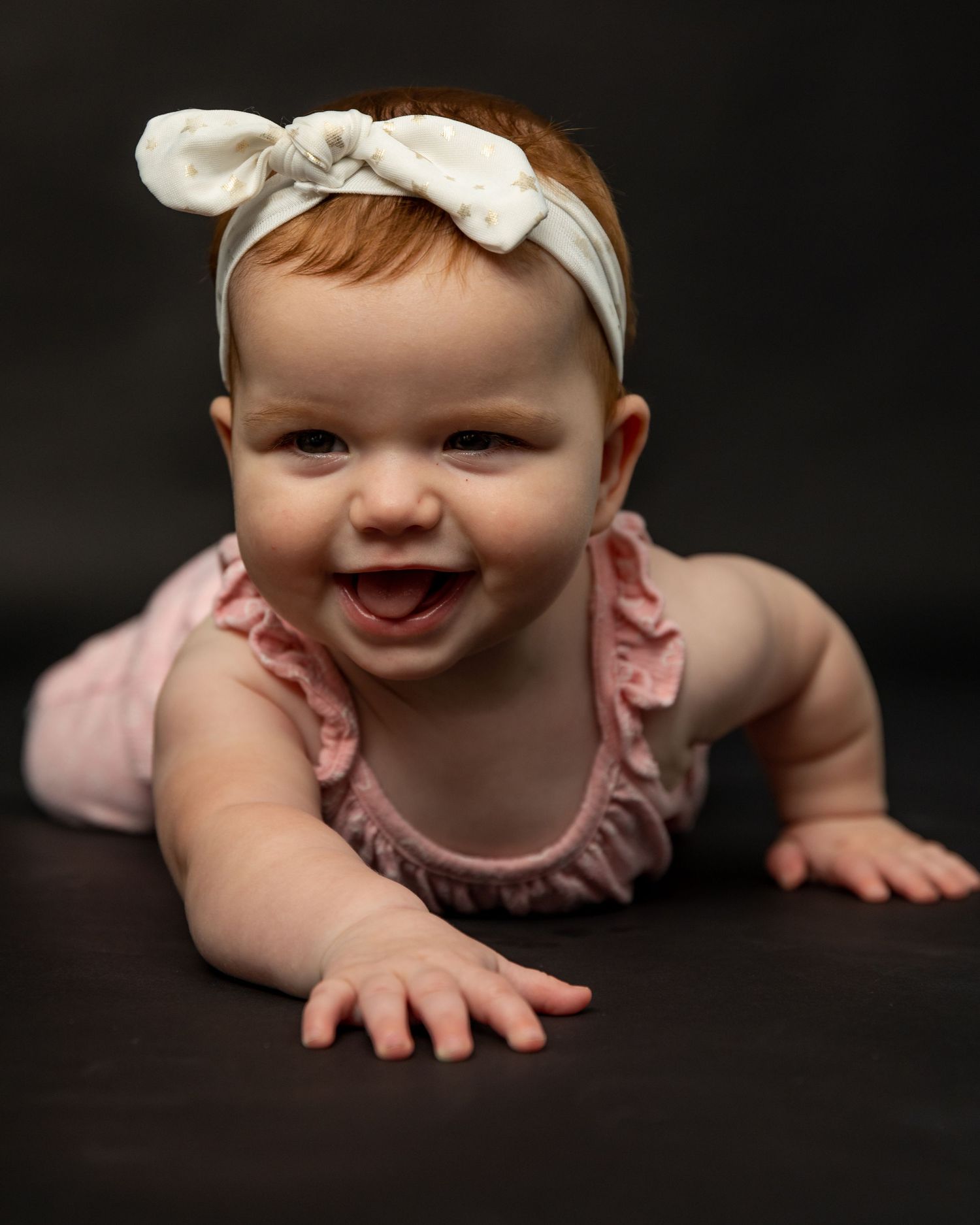 Baby with bow headband smiling during dark background photo session.
