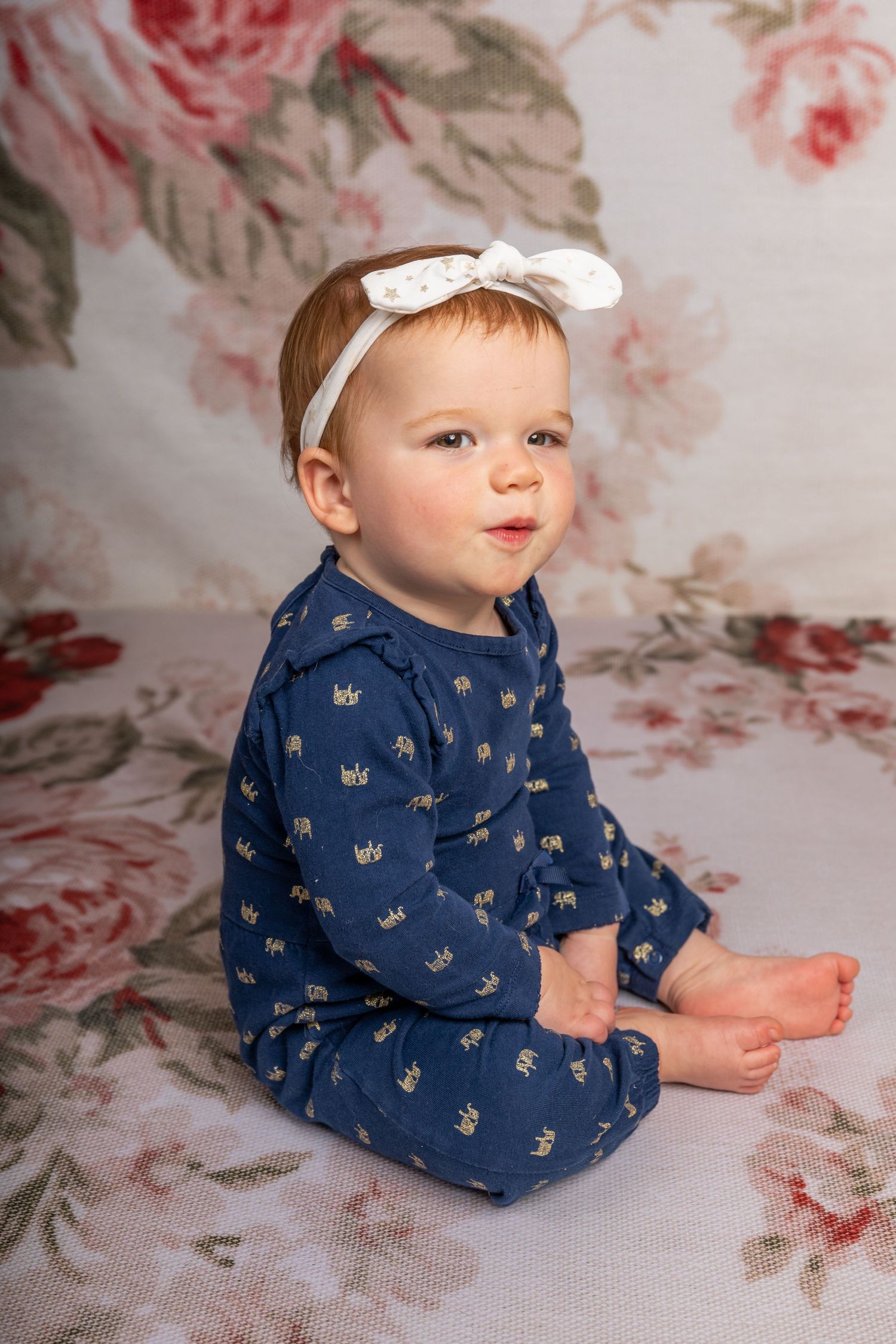 Baby wearing navy blue patterned sleepwear and white bow headband sits on floral backdrop.