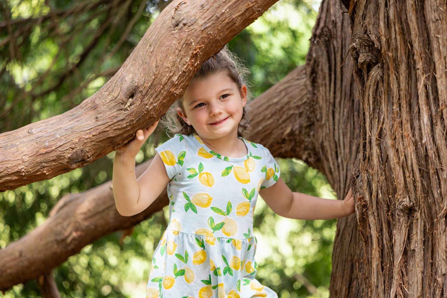Child sitting on a large tree branch in summer clothing with floral patterns.