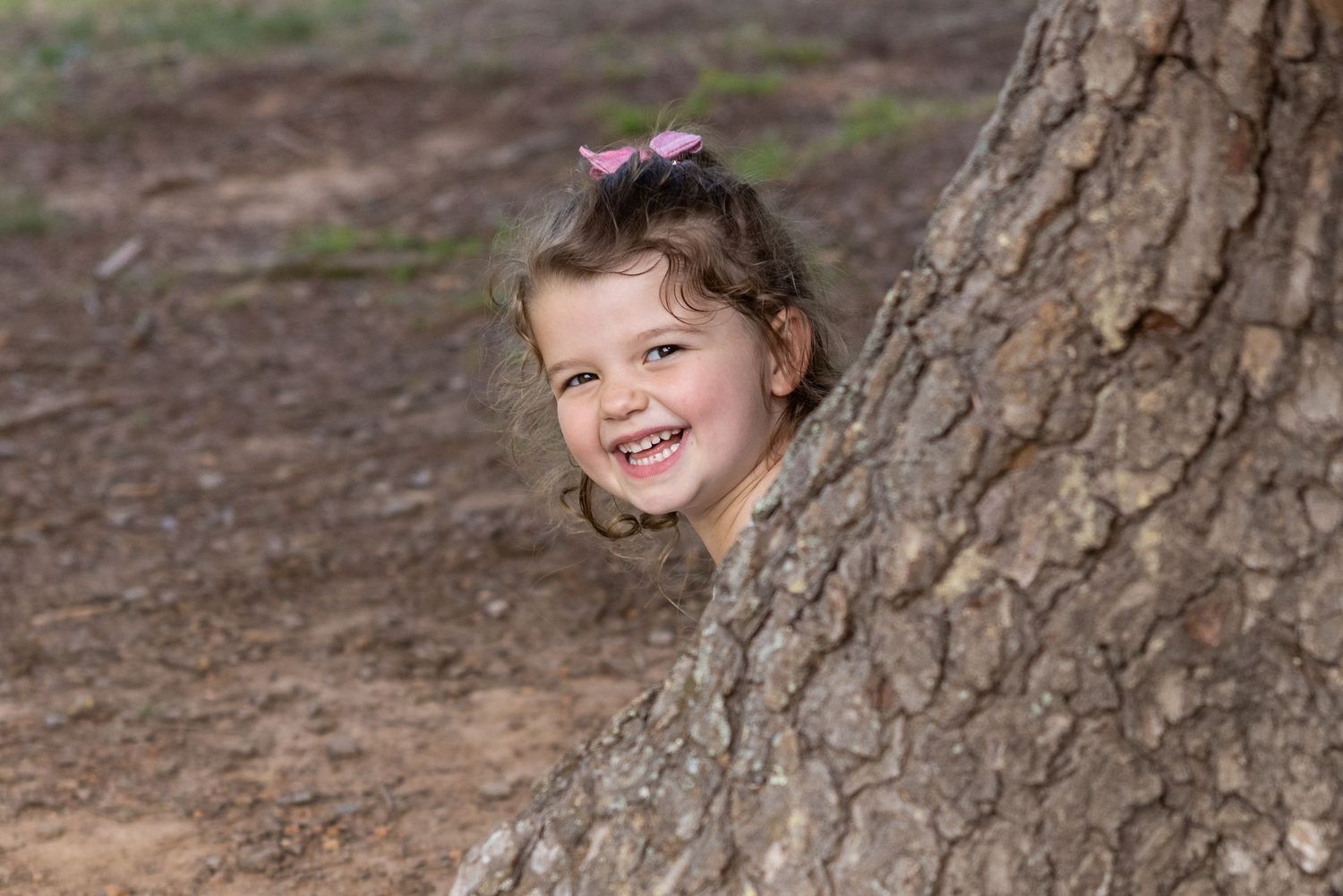 Child peeking around tree trunk with bright smile.