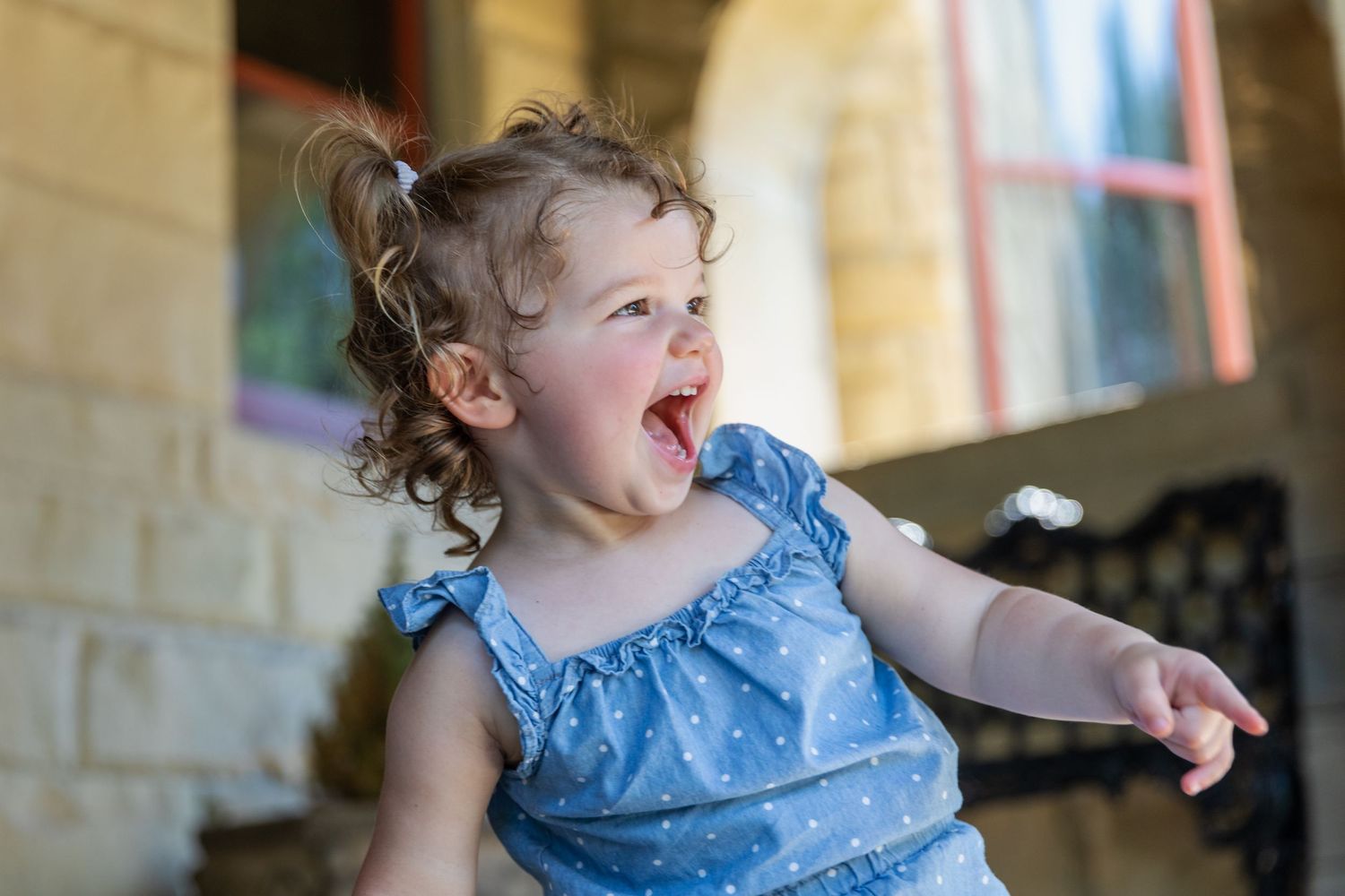 Candid moment of pure joy with toddler in blue ruffled dress against stone wall.